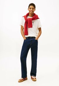 Outfit informale composto da una t-shirt bianca con grafica, jeans dritti blu scuro e una sciarpa rossa lavorata a maglia appoggiata sulle spalle.