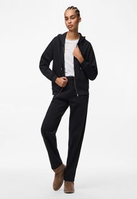 Kvinna med flätat hår iklädd en svart zip-up hoodie, vit t-shirt, svarta byxor och bruna ankelboots som står mot en enfärgad bakgrund.