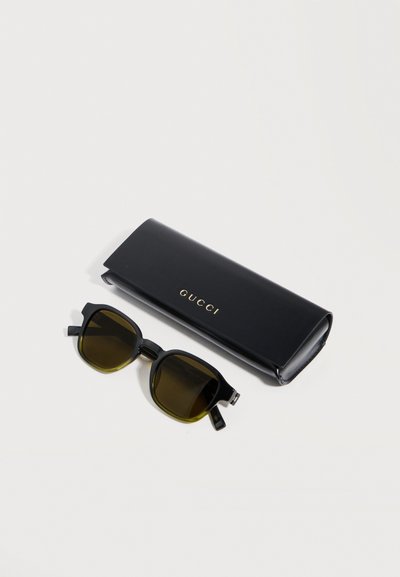 Lunettes de soleil à monture carrée noire avec des verres foncés, posées à côté d'un étui Gucci noir assorti sur un fond blanc.