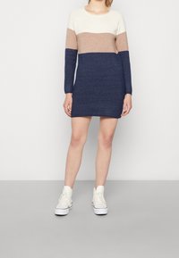 Robe-pull en blocs de couleur crème, beige et marine. Tissu en maille, manches longues, col rond et longueur mi-cuisse ; portée avec des baskets blanches.