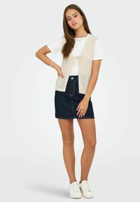 Maglione leggero beige a maglia con motivo traforato, legato in vita, sopra una maglietta bianca e una mini gonna in denim scuro, abbinato a sneaker bianche.