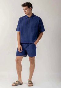 Young man wearing a blue pinstripe short-sleeve shirt with matching shorts and brown sandals, standing with one hand in pocket.