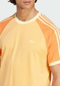 Tričko s krátkym rukávom vyrobené z mäkkého bavlneného materiálu. Obsahuje gradientný dizajn v žltej a oranžovej farbe, s bielymi tromi pruhmi a akcentmi loga.