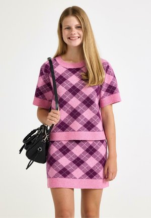 Ung kvinde smiler, iført en lyserød og lilla argylemønstret kortærmet sweater og matchende nederdel, holder en sort håndtaske.
