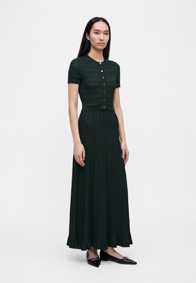 Femme portant une robe en maille vert foncé à manches courtes avec boutons, un long ourlet volanté et des ballerines noires pointues, debout sur un fond uni.