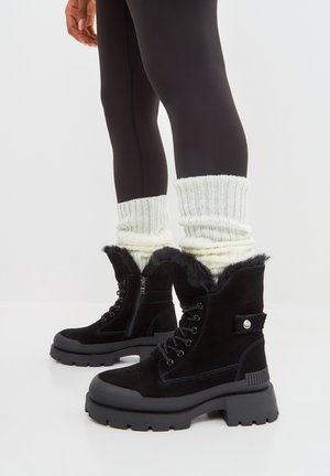 Personne portant des bottes à plateforme épaisses noires avec doublure en fourrure, des guêtres blanches en maille épaisse et des leggings noirs, debout sur un sol blanc.