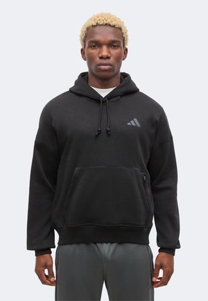 Homme aux cheveux blonds courts et bouclés, portant un sweat à capuche noir Adidas et un pantalon gris foncé, debout devant un fond clair uni.