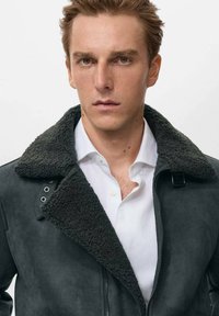 Giacca in suede grigio scuro con colletto in shearling grigio scuro testurizzato, chiusura a zip e dettagli in metallo, indossata sopra una camicia bianca.