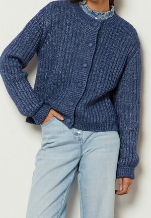 Femme portant un cardigan à boutons en maille côtelée bleue sur un chemisier à volants, associé à un jean taille haute bleu clair.