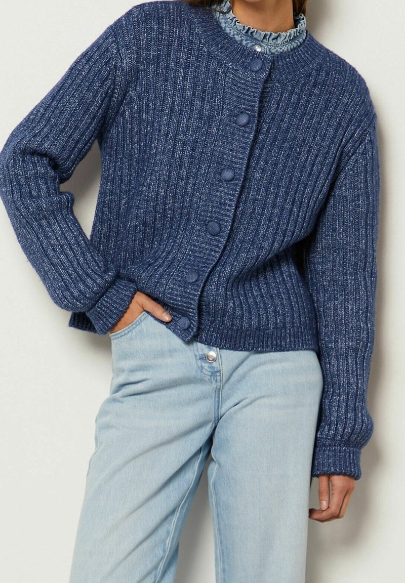 Femme portant un cardigan à boutons en maille côtelée bleue sur un chemisier à volants, associé à un jean taille haute bleu clair.