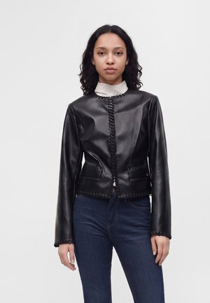 Femme aux cheveux bouclés foncés portant une veste en cuir noire par-dessus un haut blanc et un jean bleu, debout sur un fond clair uni.
