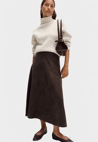 Roomkleurige ribgebreide coltrui, donkergroene suède midi-rok, zwarte platte schoenen met strikjes en een kleine bruine schoudertas. Soepele texturen.