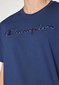 Tmavě modré bavlněné tričko s kulatým výstřihem; má vyšívané logo „Champion“ v kontrastní tmavé barvě. Měkká textura.