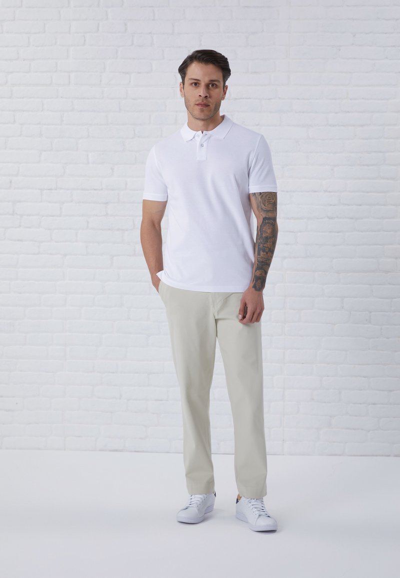 Camisa polo blanca de mangas cortas, combinada con pantalones beiges claros y zapatillas blancas. Textura suave, diseño casual.