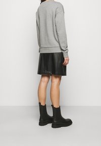 Osoba oblečená v šedé mikině, černé kožené sukni a černých kotníkových botách, stojící proti jednobarevnému světlému pozadí, otočená zády.