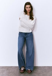 Joven mujer con cabello largo y castaño que lleva puesto un suéter blanco de punto, pantalones vaqueros de pierna ancha azul y zapatillas color burdeos, de pie contra una pared blanca.