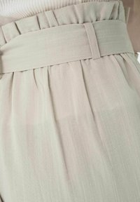 Un pantalon plissé vert clair présente une taille large avec passants de ceinture, fabriqué dans un tissu doux et texturé. Le design est décontracté et fluide.