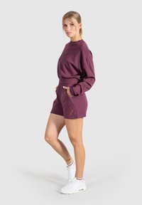 Completo felpa e pantaloni corti color bordeaux in tessuto morbido, con vestibilità ampia, vita elasticizzata e tasche. Accentato con dettagli del logo.