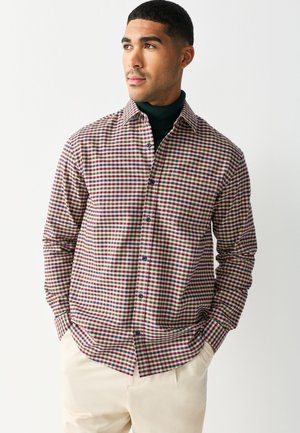 Mann trägt ein mehrfarbig kariertes Hemd über einem dunkelgrünen Rollkragenpullover mit cremefarbenen Hosen, Hände in den Taschen.