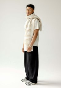 Beige textuur sjaal valt over een licht shirt. Zwarte geplooide broek en sneakers met zilveren accenten maken de outfit compleet, tegen een neutrale achtergrond.