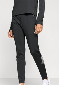 Čierne športové nohavice z hladkého materiálu. Na ľavej nohe majú biely dizajn s tromi pruhmi a bočné vrecká so zipsom.
