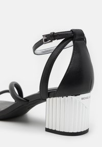 Sandale en cuir noir avec bride à la cheville, boucle argentée et talon bloc métallique côtelé de la marque « Michael Kors ».