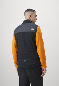 Black and orange fleece vest with high collar, featuring a logo on the back, textured surface, and contrasting sleeves. Minimalist design, zippered pockets.
