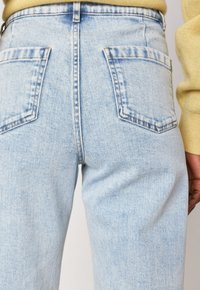 Jeans in denim azzurro chiaro con tasche posteriori indossati da una persona in un maglione giallo, mostrati dalla vita fino a metà coscia.