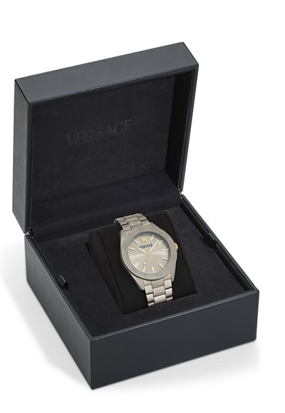 Montre Versace dans une boîte noire avec un couvercle texturé. Bracelet et boîtier en métal argenté, cadran gris avec des accents dorés et logo. Coussin noir à l'intérieur.