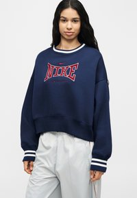 Mujer joven con cabello largo y oscuro, vistiendo un sudadera Nike azul marino con logo rojo y puños con rayas blancas, combinada con pantalones gris claro.