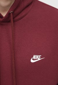 Bordová mikina vyrobená z mäkkej tkaniny s kloktanou kapsou, so bielym vyšitým logom Nike na hrudi a kapucňou so sťahovacou šnúrkou.