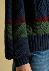 Pletený sveter s modrým telom, s horizontálnymi pruhmi v zelenej a burgundskej farbe a detailmi v káblovom pletení na rukávoch a leme.