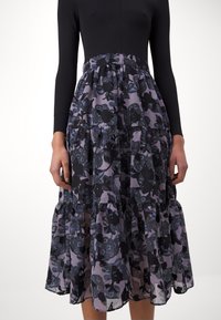 Femme portant un haut noir à manches longues et une jupe midi violette avec des motifs floraux noirs et un design à volants.