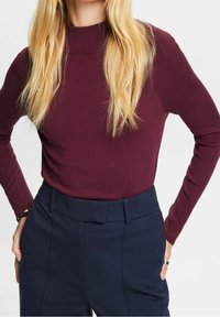 Bordowy prążkowany sweter z wysokim kołnierzem, zestawiony z ciemnogranatowymi spodniami z usztywnionym paskiem i kieszeniami na bokach.
