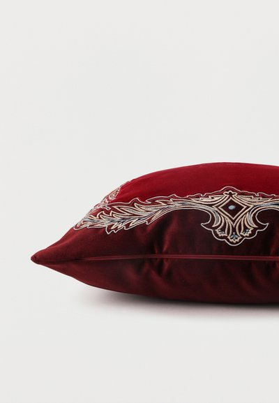 Coussin décoratif rectangulaire rouge foncé avec des motifs floraux et géométriques brodés complexes en beige et bleu clair sur un côté.