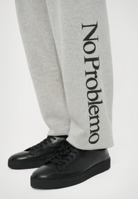 Pantalones de chándal grises con "No Problemo" impreso en negro a lo largo del costado, combinados con zapatillas de cuero negras con un acabado liso y suela plana.