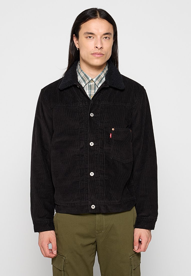 Chaqueta de pana negra con un acabado texturizado, que presenta un cuello peludo, botones en la parte delantera y un bolsillo en el pecho. Llevada sobre una camisa de cuadros.