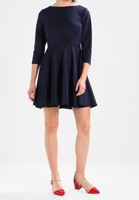 Marinblå klänning med figurnära midja och kjol, ¾-långa ärmar och rundad halsringning. Modellen bär nudefärgade klackar med ankelrem och röda detaljeringar vid tårna.