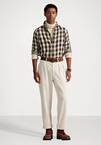 Καφέ και κρεμ καρό πουκάμισο, κρεμ ζιβάγκο, κρεμ πλισέ παντελόνι, καφέ ζώνη, καφέ και κόκκινα αρβυλάκια με κορδόνια, απλό φόντο.