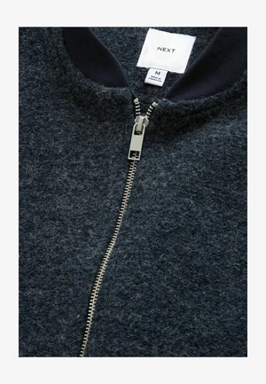 Jachetă navy blue cu fermoar, realizată dintr-un material texturat, având un fermoar argintiu și un guler cu nervuri. Eticheta cu marker indică mărimea M.