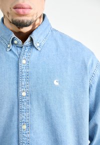 Lichtblauwe denim overhemd met knopen en een kraag, met een wit logo op de linkerborst en drie knopen gedeeltelijk vastgemaakt bij de nek.