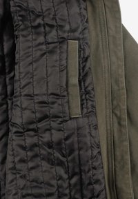 Wnętrze kurtki ocieplanej z pikowaną czarną podszewką i kontrastującym zielonym zewnętrznym materiałem. Posiada małą pętelkę z tkaniny do zawieszania.