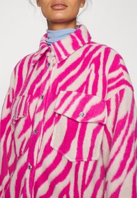 En rosa och vit zebrarandig överdimensionerad skjortjacka i mjukt tyg. Har två bröstfickor och tryckknappar för stängning.