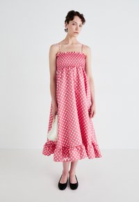Pink polka dot dress with a smocked bodice, spaghetti straps, and a ruffled hem. Model holds a white textured handbag and wears black flats.