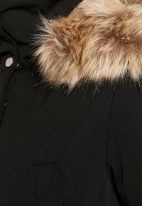 Manteau noir avec un col en fausse fourrure marron douceur, présentant une texture lisse et une poche avant sur le c ôté gauche. Détail de fermeture à pression argentée.