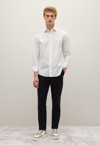 Camicetta bianca a righe con maniche lunghe, abbinata a un pantalone blu navy e sneakers chiare con suola spessa.