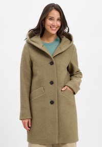 Junge Frau lächelt, trägt einen geknöpften olivgrünen Kapuzenmantel mit Händen in den Taschen, über einem türkisfarbenen Pullover und beigen Hosen.