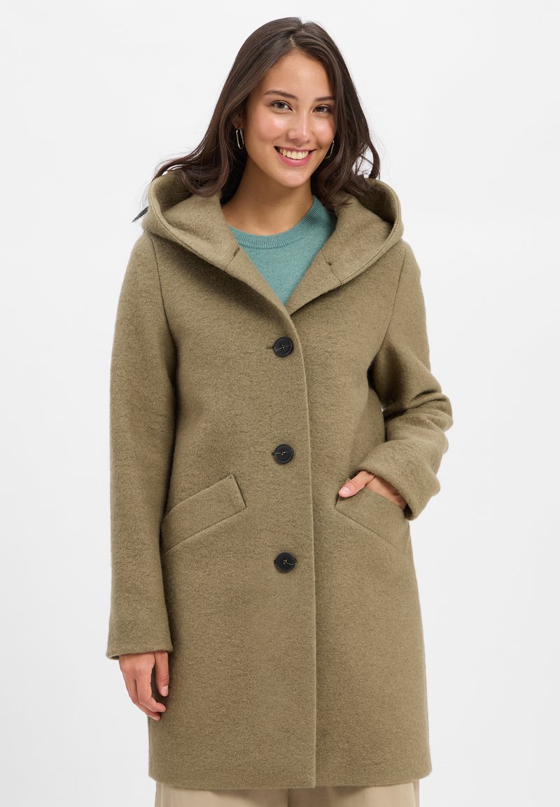 Junge Frau lächelt, trägt einen geknöpften olivgrünen Kapuzenmantel mit Händen in den Taschen, über einem türkisfarbenen Pullover und beigen Hosen.