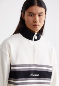 Witte sweatshirt met een hoge zwarte kraag, half-rits en horizontale strepen in grijs en zwart. Bevat het logo "ellesse" op de voorkant.