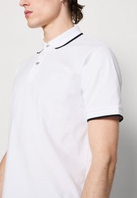 Weißes Polohemd aus Baumwolle mit einem dezenten Logo, zwei Knöpfen und schwarzem Besatz am Kragen und an den Ärmelbündchen.
